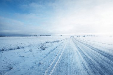 Gün batımında kar fırtınasından sonra tarlalar boyunca karla kaplı köy yolu. Arka planda eski bir köy evi var. Kırsal kesimde kış manzarası. Dramatik gökyüzü, renkli bulutlar. Buz çölü. Laponya, Finlandiya