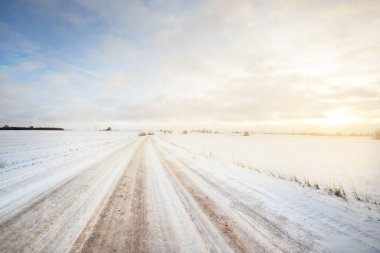 Gün batımında kar fırtınasından sonra tarlalar boyunca karla kaplı S biçimli köy yolu. Arka planda eski bir köy evi var. Kırsal kesimde kış manzarası. Dramatik gökyüzü, renkli bulutlar. Buz çölü. Laponya, Finlandiya