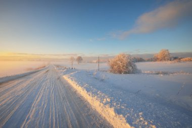Gün batımında kar fırtınasından sonra karla kaplı arazide köy yolu. Gökyüzü açık, altın ışık. Idyllic kırsal kesim. Panoramik manzara. Noel, lojistik, tehlikeli sürüş, off-road, ulaşım