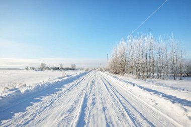 Güneşli bir günde kırsal alanda karla kaplı tek şeritli yol. Finlandiya. Açık mavi gökyüzü. Yaprak döken ve yemyeşil ağaçlar arka planda kırağı kaplamış. Idyllic kış manzarası