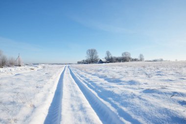 Güneşli bir günde kırsal alanda karla kaplı tek şeritli yol. Finlandiya. Açık mavi gökyüzü. Yaprak döken ve yemyeşil ağaçlar arka planda kırağı kaplamış. Idyllic kış manzarası