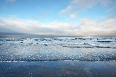 Gün batımında karla kaplı Baltık deniz kıyısından bir manzara. Riga Körfezi, Letonya. Dramatik bir gökyüzü. Kristal berrak mavi su. Kararsız hava, dalgalar ve su sıçramaları. Kış turizmi, küresel ısınma teması