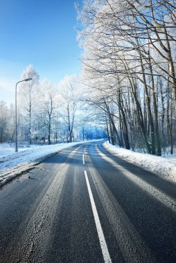 Temizlikten sonra keskin virajlı boş bir asfalt yol. Sokak fenerleri yakın plan. Taze karda araba izleri. Arka planda karla kaplı orman ve tarla var. Açık mavi gökyüzü. Kış yolculuğu. Finlandiya