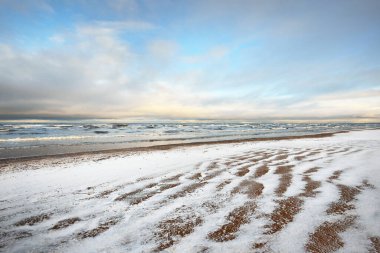 Gün batımında karla kaplı Baltık deniz kıyısından bir manzara. Riga Körfezi, Letonya. Renkli dramatik bulut manzarası. Kararsız hava, dalgalar ve su sıçramaları. Kış turizmi, küresel ısınma teması