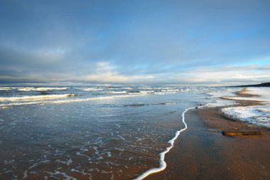 Gün batımında karla kaplı Baltık deniz kıyısından bir manzara. Riga Körfezi, Letonya. Dramatik mavi gökyüzü. Kararsız hava, dalgalar ve su sıçramaları. Kış turizmi, küresel ısınma teması