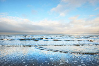 Gün batımında karla kaplı Baltık deniz kıyısından bir manzara. Riga Körfezi, Letonya. Dramatik bir gökyüzü. Kristal berrak mavi su. Kararsız hava, dalgalar ve su sıçramaları. Kış turizmi, küresel ısınma teması
