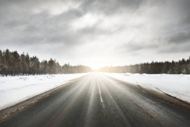 Kar kaplı tarlaların ve kozalaklı ormanların arasında kar fırtınasından sonra boş, temiz bir otoyol. Parlak bulutlu dramatik bir gökyüzü. Karelia, Rusya. Tehlikeli sürüş, yolculuk kavramları