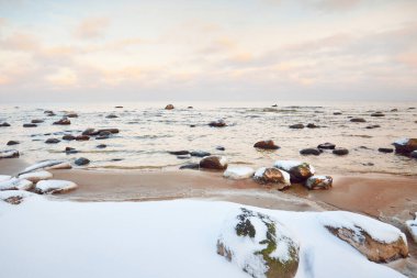 Gün batımında karla kaplı Baltık sahili manzarası. Taşlar suya yakın planda. Açık mavi gökyüzü ve renkli bulutlar. Şaşırtıcı bulut manzarası. Ilık akşam ışığı. Idyllic kış sahnesi. Kaltene, Letonya