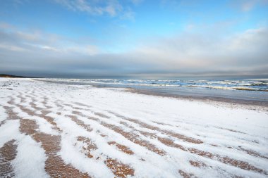 Gün batımında karla kaplı Baltık deniz kıyısından bir manzara. Riga Körfezi, Letonya. Renkli dramatik bulut manzarası. Kararsız hava, dalgalar ve su sıçramaları. Kış turizmi, küresel ısınma teması