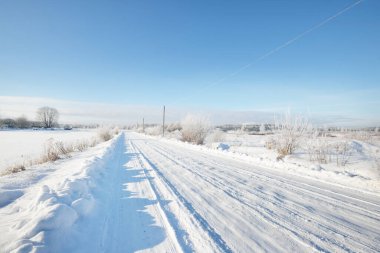 Güneşli bir günde kırsal alanda karla kaplı tek şeritli yol. Finlandiya. Açık mavi gökyüzü. Yaprak döken ve yemyeşil ağaçlar arka planda kırağı kaplamış. Idyllic kış manzarası