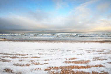 Gün batımında karla kaplı Baltık deniz kıyısından bir manzara. Riga Körfezi, Letonya. Renkli dramatik bulut manzarası. Kararsız hava, dalgalar ve su sıçramaları. Kış turizmi, küresel ısınma teması