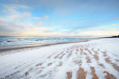 Gün batımında karla kaplı Baltık deniz kıyısından bir manzara. Riga Körfezi, Letonya. Renkli dramatik bulut manzarası. Kararsız hava, dalgalar ve su sıçramaları. Kış turizmi, küresel ısınma teması