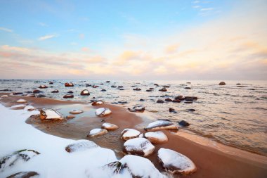 Gün batımında karla kaplı Baltık sahili manzarası. Taşlar suya yakın planda. Açık mavi gökyüzü ve renkli bulutlar. Şaşırtıcı bulut manzarası. Ilık akşam ışığı. Idyllic kış sahnesi. Kaltene, Letonya