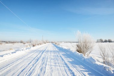 Güneşli bir günde kırsal alanda karla kaplı tek şeritli yol. Finlandiya. Açık mavi gökyüzü. Yaprak döken ve yemyeşil ağaçlar arka planda kırağı kaplamış. Idyllic kış manzarası