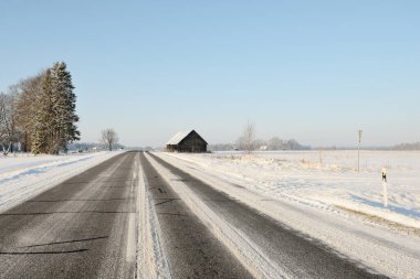 Temizlikten sonra boş bir asfalt yol. Eski bir geleneksel ev (ahşap kulübe) yakın plan. Taze karda araba izleri. Arka planda karla kaplı orman ve tarla var. Açık mavi gökyüzü. Kış yolculuğu. Letonya