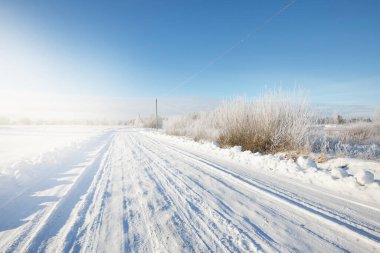 Güneşli bir günde kırsal alanda karla kaplı tek şeritli yol. Finlandiya. Açık mavi gökyüzü. Yaprak döken ve yemyeşil ağaçlar arka planda kırağı kaplamış. Idyllic kış manzarası