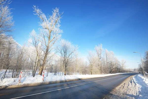 An empty asphalt road after cleaning. Street lanterns close-up. Car tracks in a fresh snow. Snow-covered birch forest in the background. Clear blue sky. Winter driving in Finland. Global warming theme