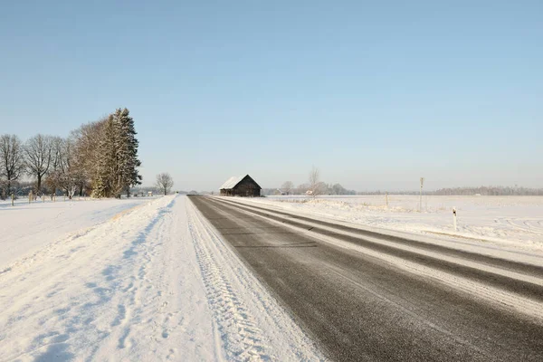 洗浄後の空のアスファルトの道路 古い伝統的な家 ログキャビン のクローズアップ 新鮮な雪の中で車のトラック 背景に雪に覆われた森とフィールド 真っ青な空 冬の運転 ラトビア — ストック写真