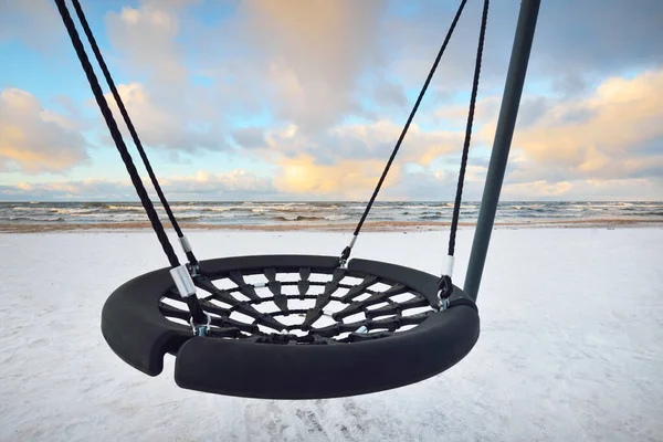Modern swing on the snow-covered Baltic sea shore at sunset. Riga bay, Latvia. Colorful dramatic cloudscape. Deep cyclone, rough weather. Winter vacations, global warming theme
