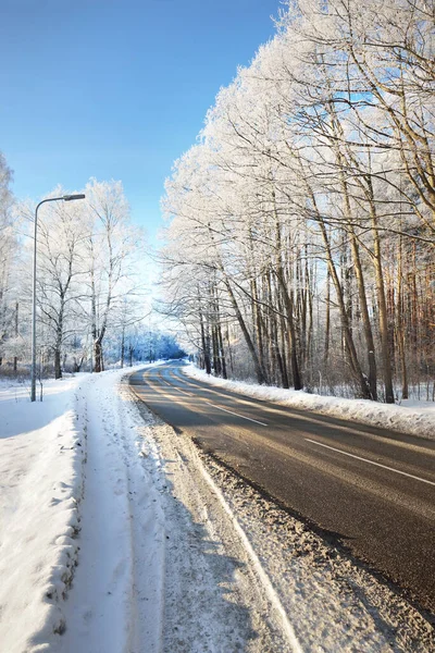 An empty asphalt road after cleaning. Street lanterns close-up. Car tracks in a fresh snow. Snow-covered birch forest in the background. Clear blue sky. Winter driving in Finland. Global warming theme