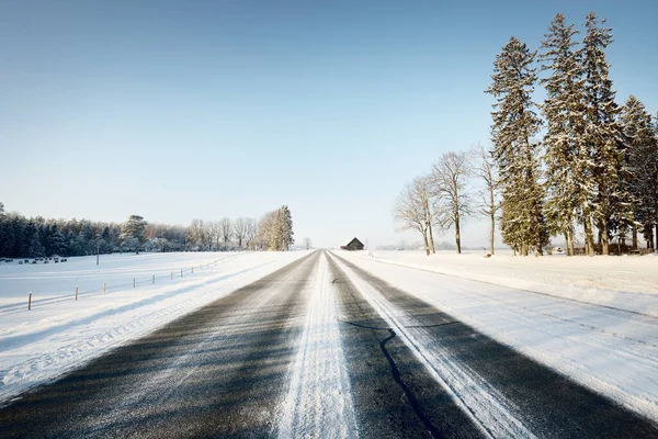 An empty asphalt road after cleaning. An old traditional house (log cabin) close-up. Car tracks in a fresh snow. Snow-covered forest and field in the background. Clear blue sky. Winter driving. Latvia
