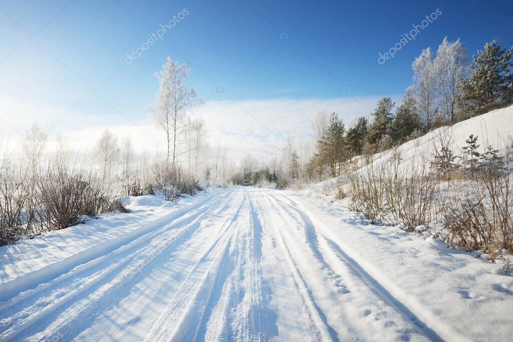 Snow-covered single lane rural road through the fields on a sunny day. Finland. Clear blue sky. Deciduous and evergreen trees in hoarfrost in the background. Idyllic winter landscape