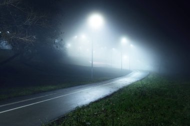 Gece sisli bir havada keskin bir dönüş yapan boş, aydınlık bir bisiklet yolu. Fenerler yakın plan. Köprü arka planda. Eğlence ve sağlıklı yaşam tarzı teması. Riga, Letonya