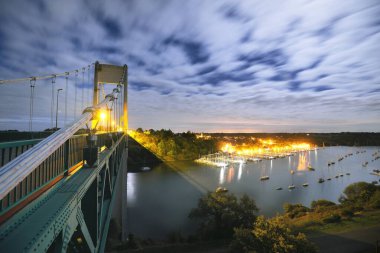 Işıklandırılmış kablo destekli yol köprüsünün panoramik hava görüntüsü. Fransa. İnşaat endüstrisi, dönüm noktası, turizm. Karanlık şehir manzarası, şehir manzarası. Arka planda yat marinası var. Uzun pozlama