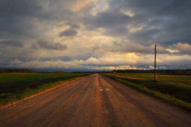 Gün batımında yağmurdan sonra tarlalardan geçen boş bir köy yolu. Dramatik renkli gökyüzü, destansı bulutlar. Transformatör direkleri yakın çekim. Letonya