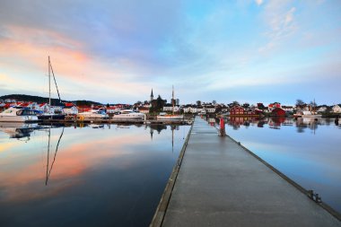 Yelkenli ve balıkçı tekneleri gün batımında yat limanındaki bir rıhtıma demirlediler. Renkli bulutlar, suda yansımalar. Yelkencilik, eğlence teması. Güney Norveç Köyü