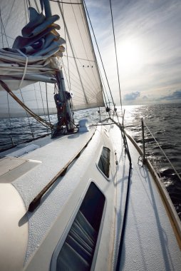 Güneşli bir yaz gününde açık denizde yelken açan beyaz bir yat. Güverteden pruva ve yelkenlere kadar yakın çekim. Dalgalar ve su sıçramaları. Sirus bulutları ve uçak izleri olan açık mavi bir gökyüzü. Hollanda