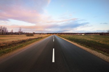 Boş bir otoyol (asfalt yol) yeşil kırsal tarım tarlaları gün batımında sürüldü. Güzel pembe bulutlar. Sıcak kış. İklim değişikliği teması. Letonya