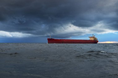Büyük kırmızı kargo gemisi karanlık günbatımı göğünün altında açık denizde yol alıyor. Southampton 'da. İngiltere. Dramatik bulutlar, kasırga, fırtınalı hava. Küresel iletişim, nakliye, endüstri, lojistik