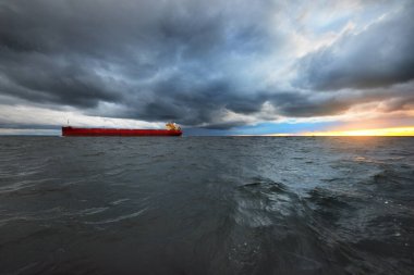Büyük kırmızı kargo gemisi karanlık günbatımı göğünün altında açık denizde yol alıyor. Southampton 'da. İngiltere. Dramatik bulutlar, kasırga, fırtınalı hava. Küresel iletişim, nakliye, endüstri, lojistik