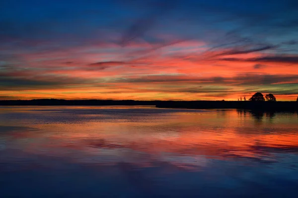 Epic red sunset sky above the lake and forest in autumn. Dramatic cloudscape. Symmetry reflections on the water, natural mirror. Idyllic rural scene. Gauja national park, Sigulda, Latvia