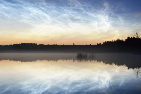 Starry sky with noctilucent clouds and fog above the Saimaa lake, Finland, at summer solstice night. Long exposure. Symmetry reflections, natural mirror. Mystic landscape, pagan celebration