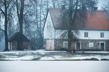 Kırmızı kiremitli ve eski yosunlu ağaçlı geleneksel kır evi. Bulutlu bir günde kar fırtınası. Kırsal kesimde kış manzarası. Almanya