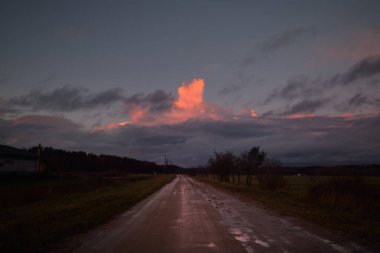 Gün batımında yağmurdan sonra tarlalarda boş bir kırsal yol. Dramatik renkli gökyüzü, destansı bulutlar. Letonya