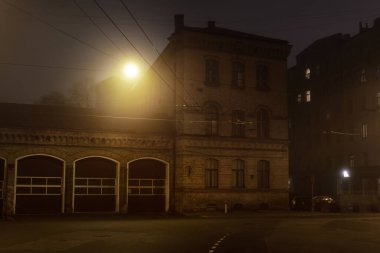 Geceleyin sisli bir şekilde eski tarihi binaların ve evlerin arasından aydınlatılmış boş bir asfalt yol. Sokak lambaları (fenerler) yakın plan. Riga, Letonya. Karanlık şehir manzarası