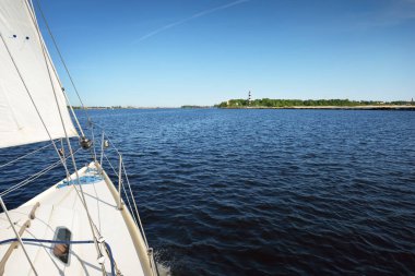 Açık bir yaz gününde deniz fenerinin yanında yelken açan beyaz bir yat. Güverteden pruva ve yelkenlere kadar yukarıdan aşağıya doğru. Ruhnu Adası, Estonya