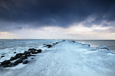 Boş gezinti güvertesi ve karla kaplı dalgalar yakın plan, arkada donmuş Baltık denizi. Kara fırtına bulutları. Kış, mevsimler, iklim değişikliği, küresel ısınma kavramları. Uzun pozlama