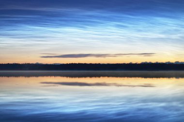 Yaz gündönümü gecesinde Saimaa Gölü 'nün üzerinde gece parlayan bulutlar ve sis vardı. Ağaç siluetleri. Altın güneş ışığı. Uzun pozlama. Destansı bulut manzarası. Suyun simetri yansımaları. Finlandiya