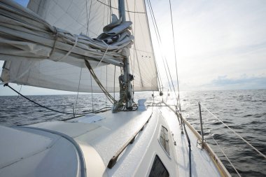 Güneşli bir yaz gününde açık denizde yelken açan beyaz bir yat. Güverteden pruva ve yelkenlere kadar yakın çekim. Dalgalar ve su sıçramaları. Sirus bulutları ve uçak izleri olan açık mavi bir gökyüzü. Hollanda