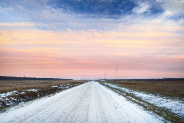 Tarlalarda karla kaplı köy yolu, trafolar yakın çekim. Arka planda orman var. Renkli bulutlarla dramatik günbatımı gökyüzü. Kış manzarası. Letonya