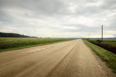Yağmurdan sonra tarlalardan geçen boş bir köy yolu. Dramatik gökyüzü, destansı bulutlar. Kötü hava. Transformatör direkleri yakın çekim. Letonya