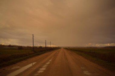 Gün batımında yağmurdan sonra tarlalardan geçen boş bir köy yolu. Dramatik renkli gökyüzü, destansı bulutlar. Transformatör direkleri yakın çekim. Letonya