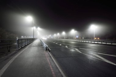 Gece sisli bir havada karayolu yakınında boş bir bisiklet yolu (yaya geçidi). Fenerler yakın plan. Ulaşım, tehlikeli sürüş, eğlence ve sağlıklı yaşam tarzı teması. Riga, Letonya