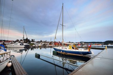 Mavi yelkenli gün batımında yat limanındaki bir rıhtıma demirlemiş. Renkli bulutlar, suda yansımalar. Yelkencilik, eğlence teması. Güney Norveç Köyü