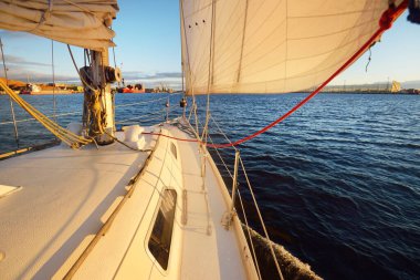 Gün batımında Daugava nehrinde beyaz bir yat. Güverteden pruvaya, direğe ve yelkenlere yakın görüntü. Arka planda yük gemileri ve iskele vinçleri var. Riga Körfezi, Letonya