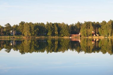 Kır evleri, yemyeşil ağaçlar ve Letonya 'da gün batımında bir nehir. Suyun simetri yansımaları. Sonbahar manzarası, huzurlu kırsal manzara. Çevre koruma, eko turizm teması